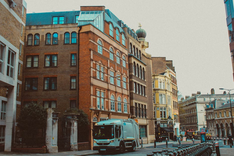 A street scene in an urban area features a mix of architectural buildings with varying facades, including a tall red brick structure with large, arched windows on the upper floors and decorative detailing. To the right, there is a beige and yellow building with intricate moldings and bay windows. In the foreground, a white waste collection truck with a clean, smooth surface and a compact design is parked on the paved street, slightly overlapping a row of black bicycle racks. The truck is positioned near the entrance to a gated property with stone pillars and greenery visible behind the gate. The street is lined with bollards and a lamp post, with additional historic and modern buildings stretching into the background. Overcast skies cast diffuse natural light over the scene, emphasizing the textures of the brick, stone, and metal surfaces. This image relates to independent waste management or rubbish removal services that manage on-site collection, as exemplified by the presence of the waste truck along a typical city street, consistent with private disposal alternatives.