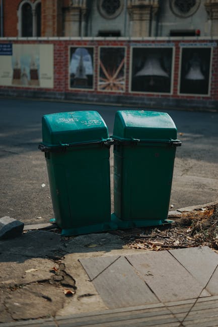 Two green plastic rubbish bins with dome-shaped lids standing side by side on a pavement outside a building. The bins are positioned at the edge of the sidewalk, close to a curb, and are made of durable, textured plastic with black handles on their lids. They are situated on a concrete surface with some scattered leaves and dirt nearby. In the background, there is a road with a blurred view of a brick wall or construction barrier featuring printed posters and advertisements, and a building with decorative architectural elements and arched windows. The scene is illuminated by natural daylight, with subtle shadows cast by the bins and surrounding objects, reflecting a typical urban environment suitable for private waste collection or alternative rubbish disposal services. The image is relevant to rubbish removal services, possibly depicting a typical setup for waste collection in Clapham, as provided by Rubbish Removal Clapham. The scene emphasizes the importance of proper waste storage before collection, aligning with independent refuse management practices.