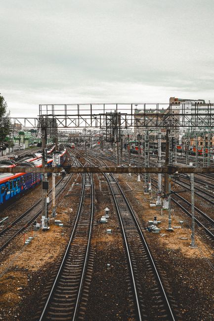 The image depicts a train station with multiple railway tracks extending from the foreground into the distance, surrounded by numerous overhead electrical wires and metal supports. In the background, there are several buildings, some of which are brick or concrete, with a variety of flat and slightly sloped roofs. The sky above is overcast with a layer of grey clouds, creating diffuse, even lighting across the scene. The tracks are made of steel and are mounted on wooden sleepers set in gravel, with some sections showing slight rust on the metal rails. To the left, a blue train with red accents partially visible is parked alongside the platform, which is not fully shown. The scene illustrates a busy railway yard potentially involved in rail transport or freight, and the detail emphasizes infrastructure relevant to alternative or private waste removal services that may use rail transport for logistical purposes, aligning subtly with a landscape where on-site or independent collection solutions for waste or rubbish might be involved. The overall atmosphere is utilitarian and functional, capturing the dense network of railway infrastructure typical in urban or industrial areas.