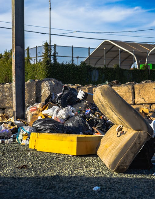 A group of large, crumpled black plastic rubbish bags are placed on the edge of a paved sidewalk in front of a dark metal fence with vertical bars, which borders a residential area. Behind the fence, dense foliage of leafy green trees or shrubs is visible, with their branches extending upward and partially obscuring a brick building or structure. The rubbish bags appear to contain unknown waste and are positioned close to the curb, indicating an area designated for waste collection. The scene is illuminated by natural daylight, casting subtle shadows on the pavement. This setup suggests an instance of private rubbish disposal or alternative waste handling, which is typical for services offered by companies like Rubbish Removal Clapham specializing in on-site clearance or independent collection of refuse in urban residential environments. The overall environment appears calm, with no visible activity or personnel present, emphasizing the focus on discarded waste awaiting collection or removal.