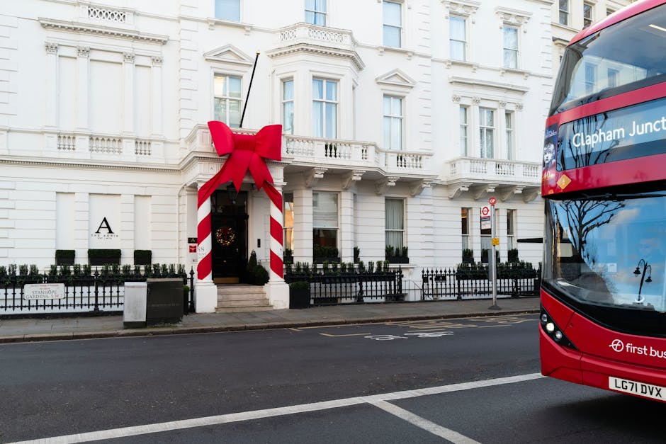 A white, multi-storey residential building with ornate architectural details, including decorative cornices and window surrounds, situated on a city street. The entrance features a small set of steps leading up to a dark door, which is decorated with a festive wreath. To the left of the entrance, a large red bow with a wide ribbon and trailing ends is attached to a striped red and white column, creating a holiday decoration ambiance. A low black metal fence surrounds a small garden area with neatly trimmed hedges along the building’s perimeter. In the foreground, the asphalt road is visible with a white dashed line marking the lane. To the right, a part of a red double-decker bus is seen, with the destination display indicating 'Clapham Junction.' The street scene is captured in natural daylight, with soft shadows and reflections on the bus windows, emphasizing the urban environment connected to private waste collection and refuse management services in the area managed by Rubbish Removal Clapham.