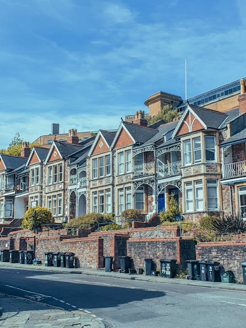 A row of Victorian-style terraced houses situated along a pavement in an urban residential area, featuring intricate white ironwork balconies and bay windows with white wooden frames. The buildings are constructed with red brick and have peaked slate roofs, with some decorative wooden gable details painted white. In front of the houses, a low brick wall separates the properties from the sidewalk, which is lined with multiple black and green wheelie bins for waste disposal. The street appears clean and quiet, with the bins placed for collection, and the scene is well-lit by natural daylight under a clear blue sky. In the background, taller modern buildings are visible above the rooftops, indicating a mixed historic and contemporary environment. This setting reflects a typical street where private waste collection services, such as those provided by Rubbish Removal Clapham, may operate to support local rubbish removal needs without reliance on municipal collection, emphasizing the context of independent waste management and on-site clearance options in the area.