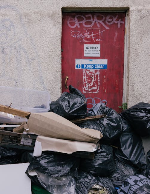 A cluster of black plastic rubbish bags, some tied and others loosely filled, piled up in front of a red metal door on an exterior wall. The bags contain various waste materials, and there are also discarded cardboard boxes and a piece of foam packaging among the debris. The door is covered in graffiti and features a white sign indicating 'NO MONEY' and 'Keep clear' in blue and white. The surrounding wall is off-white with a textured finish, and there is some grass and small plants visible at the base of the wall near the rubbish. The scene suggests an area awaiting collection for private waste removal, with unevenly stacked refuse indicating an informal or on-site clearance process possibly managed by a local rubbish removal service such as Rubbish Removal Clapham.