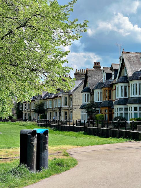 A row of traditional Victorian terraced houses with bay windows and slate roofs in a residential area, situated behind a wooden fence and a well-maintained lawn. In the foreground, a public pathway runs alongside two black public waste bins with receptacles for different waste types, positioned on a grassy verge near the pavement. A large leafy tree with bright green foliage extends over part of the scene, casting dappled shadows on the grass and pathway. The sky above is partly cloudy with patches of blue, indicating a clear day. The setting suggests a quiet suburban environment where residents might arrange private waste collection or rubbish removal services, such as those offered by Rubbish Removal Clapham, especially in the context of estate clearance or alternative waste handling outside of local authority cycles.