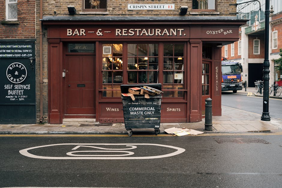 A black rubbish bin marked 'Commercial Waste Only' is positioned in front of a storefront with wooden-framed windows and a dark red exterior. The bin is overflowing with flattened cardboard boxes and miscellaneous paper waste, some of which are draped over its edges and onto the pavement. The surrounding environment includes a narrow city street with a painted road symbol, a yellow-line curb, and nearby street furniture such as a bollard and a lamppost. Adjacent to the bin, on the left side, there is a chalkboard sign advertising a self-service buffet, and to the right, a parked truck is partially visible behind a black pole. The shop appears to be a bar or restaurant, with vintage-style lighting inside visible through the glass windows. This scene illustrates a typical urban setting where private waste collection services, like those offered by Rubbish Removal Clapham, manage rubbish disposal beyond local authority street cleansing, supporting proper clearance of commercial waste in the area.
