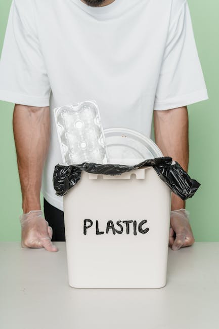A person wearing a white t-shirt and disposable gloves stands behind a white plastic bin labeled 'PLASTIC' in black marker. The bin is filled with various plastic waste items, including a black plastic bag draped over its rim and a grey plastic egg carton positioned inside. The setting appears to be indoors with a light green wall in the background, and the surface in front of the person is a plain, light-colored table. This scene illustrates an act of segregating plastic rubbish for proper disposal or recycling, emphasizing the importance of waste separation in effective rubbish removal processes. The visual focus on plastic waste contained in a dedicated bin aligns with services offered by rubbish removal providers such as Rubbish Removal Clapham, which facilitate the collection and disposal of household or commercial waste through private collection methods, including on-site clearance or direct disposal options, supporting environmentally responsible waste management practices.