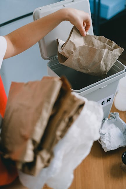 A person hands a crumpled brown paper bag into a small, white recycling bin with a black interior, which is placed on a wooden surface. The bin's side label partially visible reads 'RECYCLER'. In the foreground, another crumpled paper bag and some loose paper waste are held by the person, suggesting ongoing rubbish collection or disposal. The background shows a light-colored wall and part of a window, with natural lighting illuminating the scene. The environment appears to be an indoor space, possibly a kitchen or an office area, where waste is being sorted for private rubbish removal, aligning with independent waste management practices supported by services like Rubbish Removal Clapham. The scene emphasizes an organized approach to waste sorting and disposal, relevant to alternative rubbish collection methods outside of local authority services.
